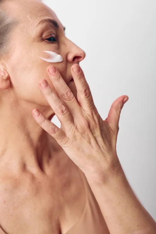 Mature woman applies face cream on cheek with fingers close-up. Skincare concept with natural skin texture, anti-aging care, beauty routine, and self-care in soft neutral lighting.