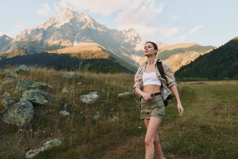 Young woman hiking outdoors in nature, wearing casual outfit with backpack, walking on grassy trail, mountain landscape background, enjoying fresh air and freedom