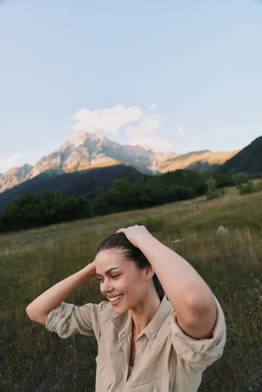 Woman enjoying outdoor lifestyle in a grassy field with distant mountains, smiling and relaxed, embodying nature, happiness, and summer vibe in a bright countryside setting