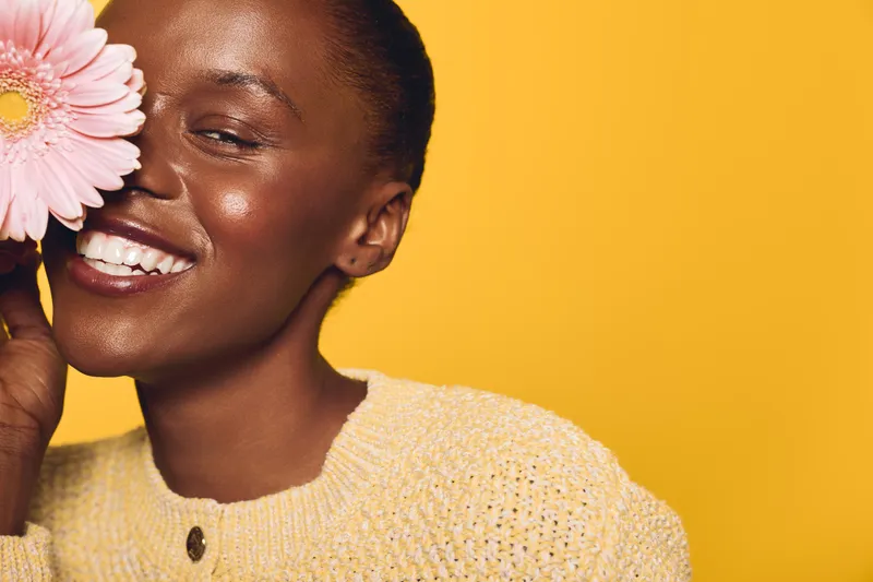 Smiling black woman with dark skin holds pink flower near face, wearing yellow sweater. Studio portrait on bright yellow background. Concept of happiness, beauty, and natural skincare.