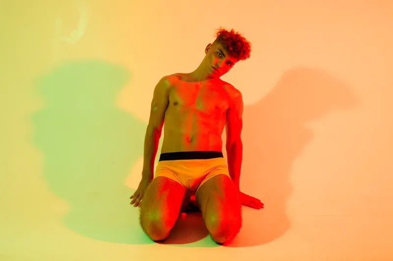 Young man with curly hair posing shirtless in colorful studio lighting, wearing white underwear, sitting on knees with artistic shadows on wall behind him.