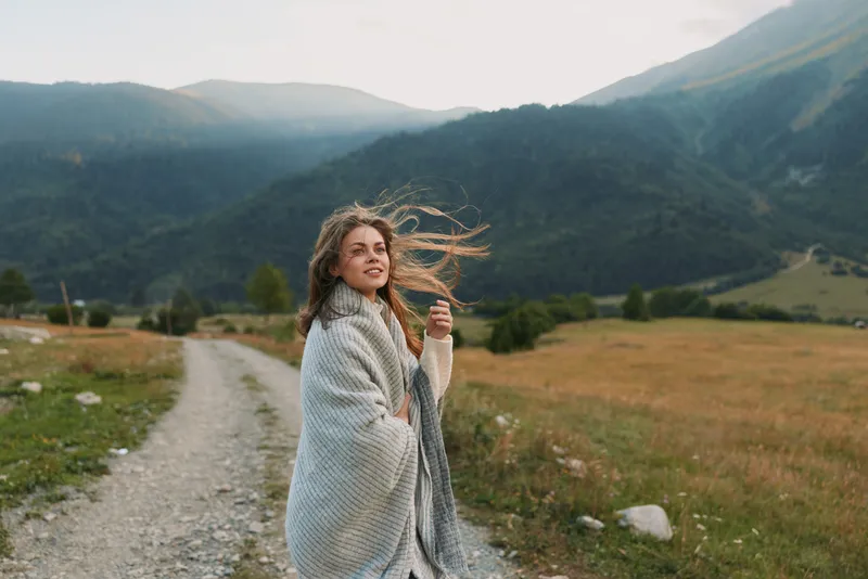 Young woman wrapped in a cozy shawl smiles as she walks along a mountain trail, wind in her hair, surrounded by a wide valley