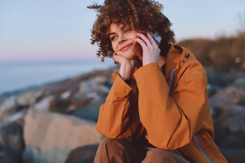 Woman with curly hair holds a phone by her ear on a rocky coast, wearing orange jacket, outdoor lifestyle near sea horizon, soft daylight, relaxed smile, communication scene today
