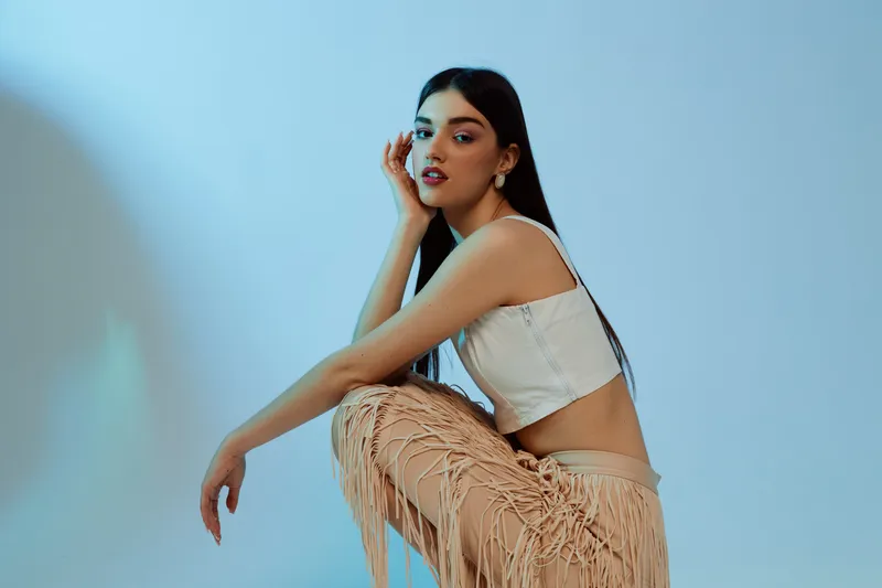 Young woman beauty fashion model posing in trendy beige fringed pants and white crop top. Studio shot with soft lighting on a blue background. Concept for style and elegance.