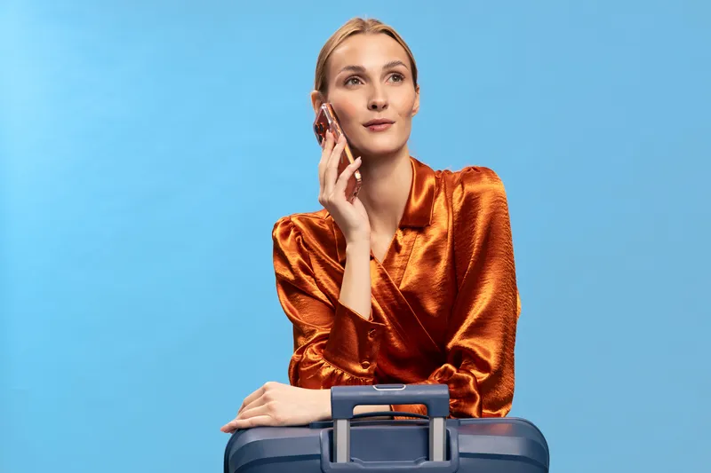 Young woman in orange satin blouse holds phone to ear and leans on blue suitcase against blue background. Concept of travel, communication, and lifestyle with modern fashion style.