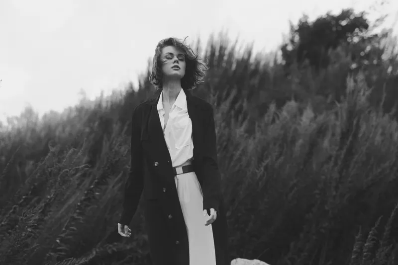 Woman in a long coat and white dress stands in tall grass, wind in hair, a moody black and white fashion shot highlighting elegance, mystery and outdoor style for editorial use and fashion