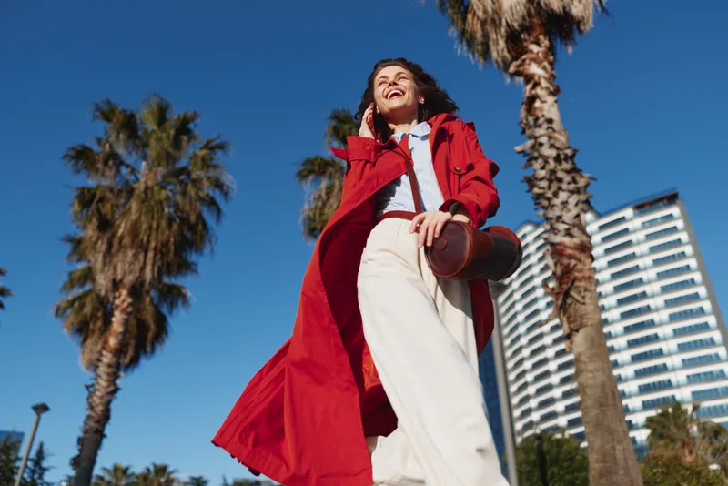 A smiling woman in a red coat talks on the phone outdoors with palm trees and a modern building in the background under a clear blue sky.