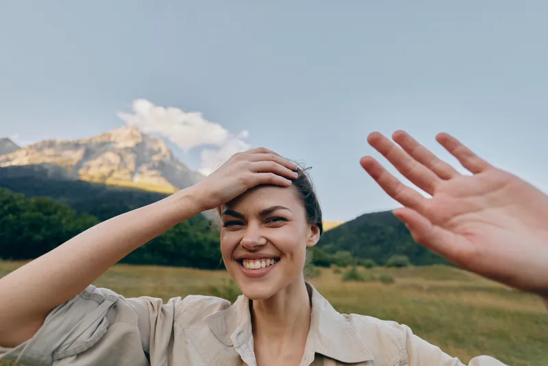 Smiling woman in casual shirt takes selfie outdoors with mountain landscape behind her, enjoying nature and lifestyle in sunny day with positive mood and relaxed expression.