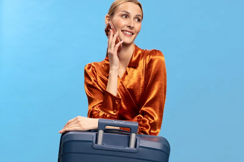 Smiling woman in shiny orange blouse with suitcase leans on luggage handle against blue background. Concept of travel, vacation, and business trip preparation with positive mood and style.