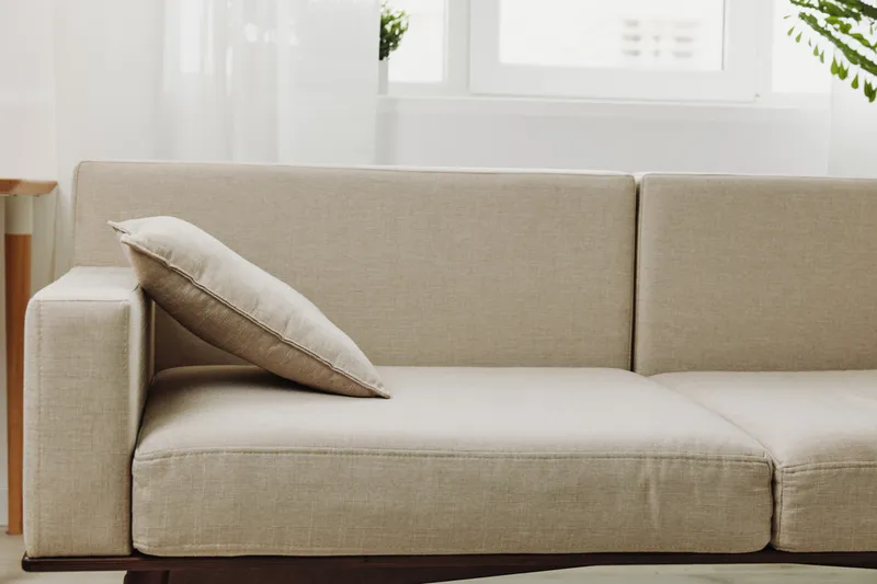 Minimalist beige fabric sofa with a single cushion in a bright living room, natural light from window, modern interior design concept for comfort and relaxation.