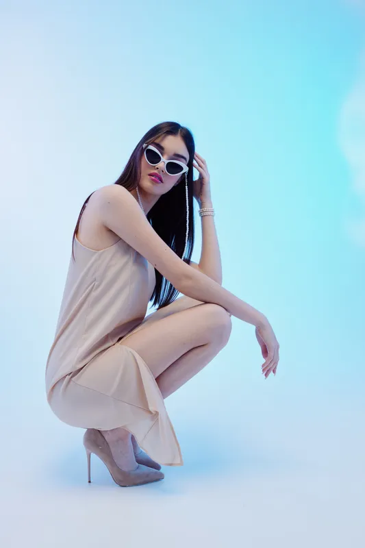 Fashionable woman with long hair wearing sunglasses and a beige dress squats on a light blue background. Concept for beauty, fashion, style, and modern elegance.