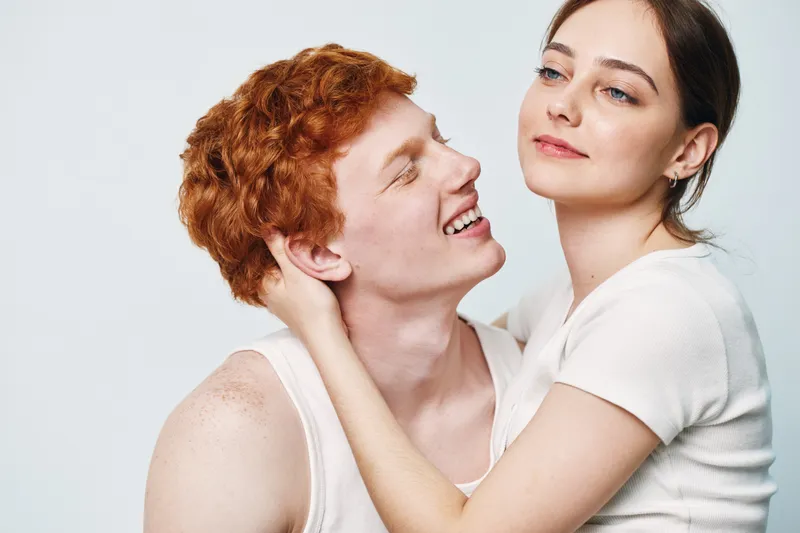 Couple, love, intimacy, romance, affection, young man with red hair and woman with white top embracing on white background with happy and thoughtful expressions.
