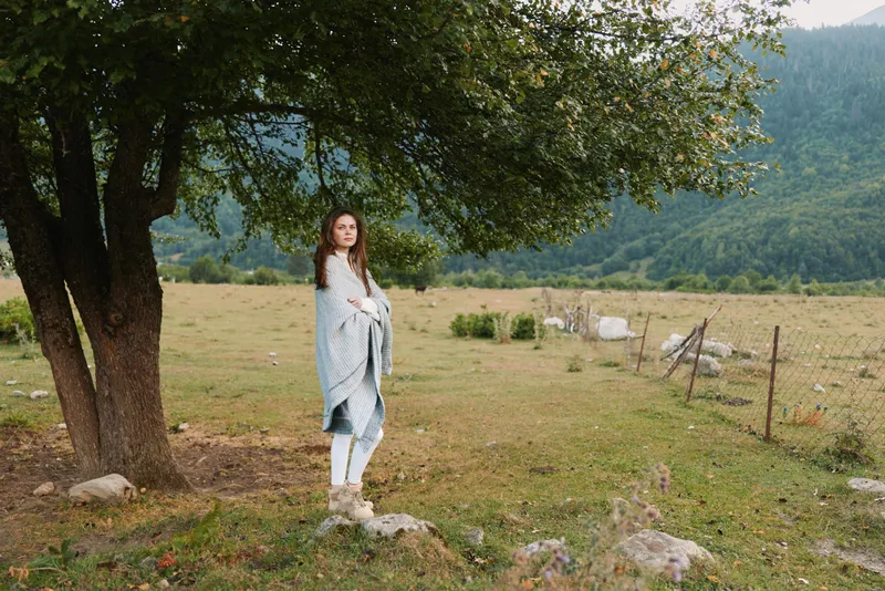 A woman wrapped in a blanket stands under a tree in a rural field with mountains in the background. The scene reflects calm lifestyle and outdoor relaxation.