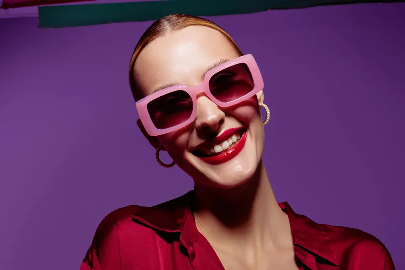 Smiling woman with pink sunglasses and red lipstick poses against purple background. Fashion portrait with bright colors and confident expression. Concept of style and beauty.