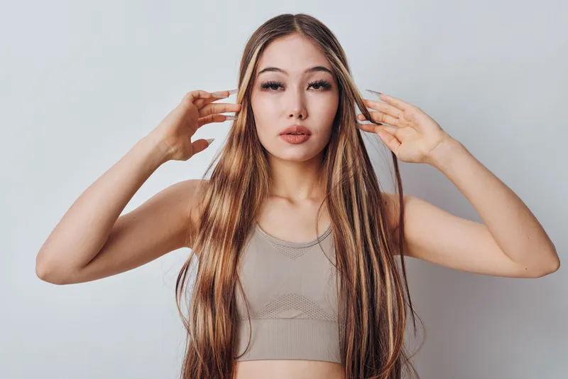 Portrait of young woman with long hair wearing crop top posing with hands near face on gray background. Female model looking at camera with natural makeup and straight hair.