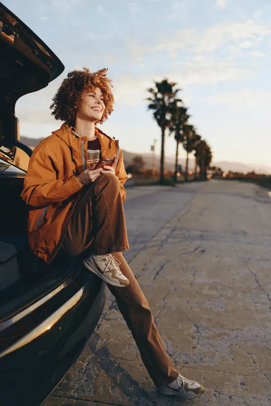 Woman lifestyle scene, smiling with curly hair while leaning on a car at golden sunset outdoors beside a quiet road, palm trees in the distance, relaxed travel mood, warm natural colors
