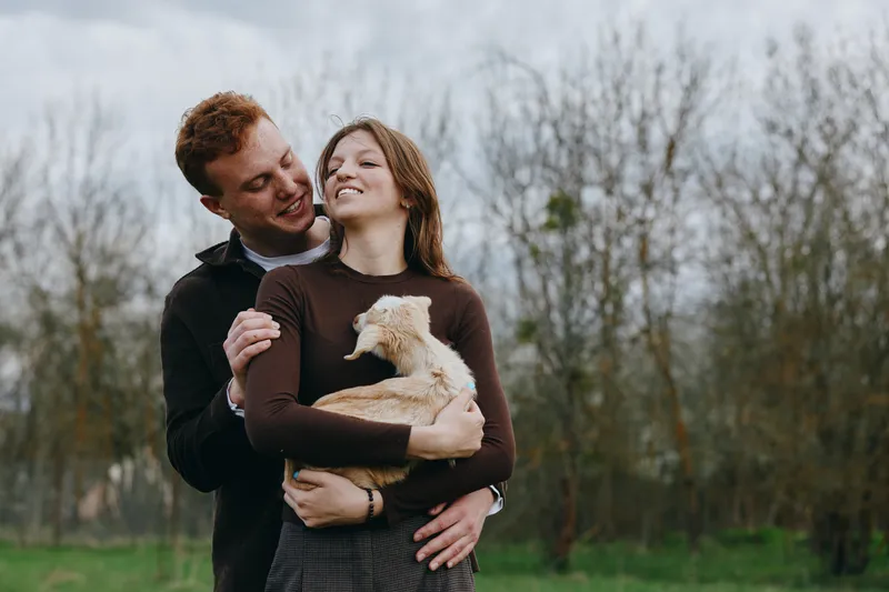 Happy couple outdoors embraces a small light dog, sharing affection in a park, warm casual mood, natural light, cheerful lifestyle moment with gentle closeness