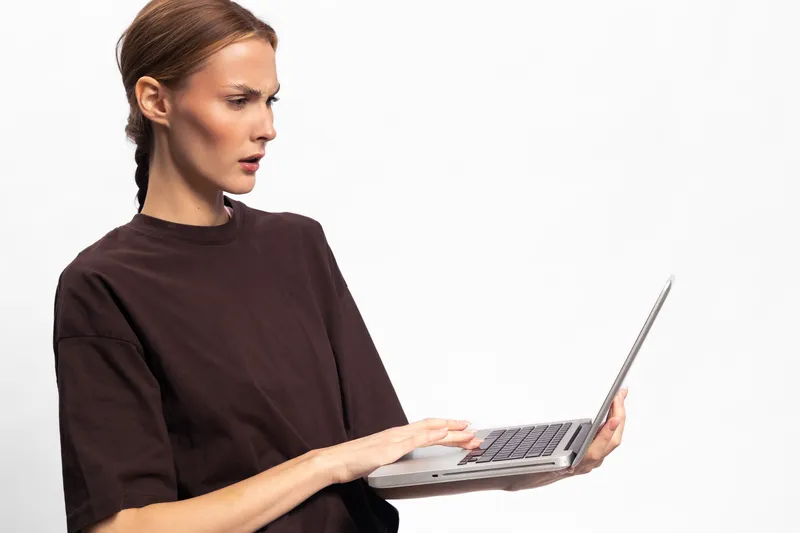 Focused woman in dark shirt holds laptop with one hand and types with the other on white background. Concept of remote work, concentration, and technology usage in modern lifestyle.