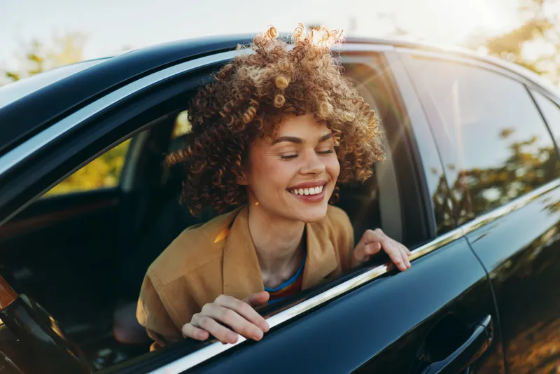 Smiling woman with curly hair leaning from a car window in a casual travel scene. Bright natural light, warm tones, and a carefree lifestyle mood. The image captures spontaneity, comfort, and modern