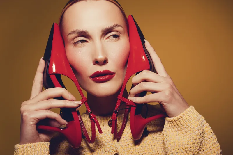 Portrait of a woman holding red high heels near her face, wearing a yellow sweater. Concept of fashion, style, and creative beauty with warm studio lighting.