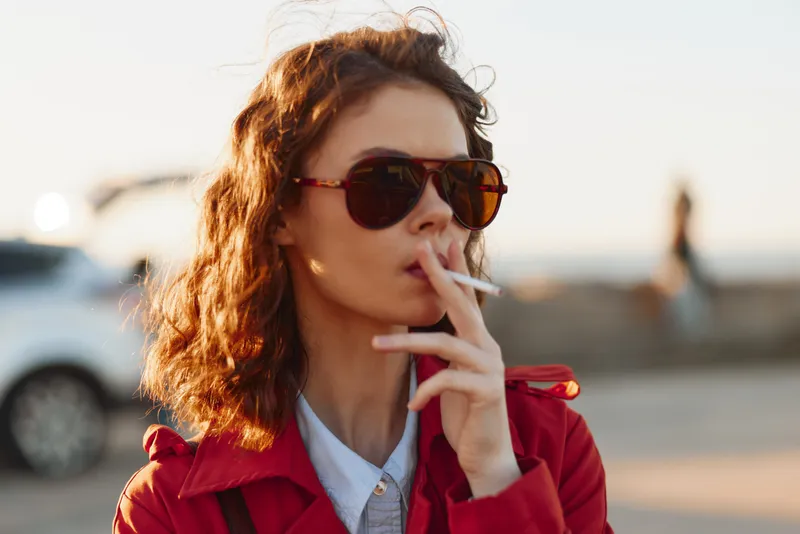 Young woman in sunglasses smoking a cigarette outdoors during golden hour. Casual style with red coat and curly hair. Urban lifestyle and relaxation concept.
