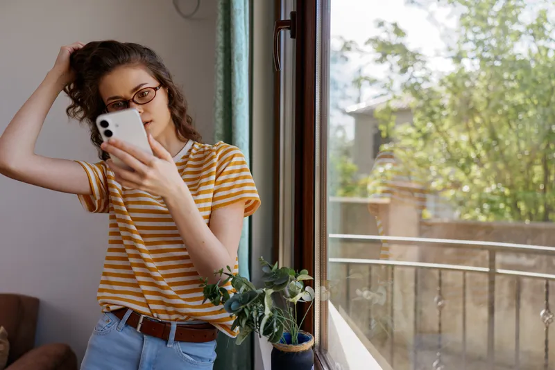 A young woman at home checks her smartphone by an open window, wearing a striped tee, relaxed pose. This scene captures everyday mood, digital lifestyle, and contemporary domestic life.