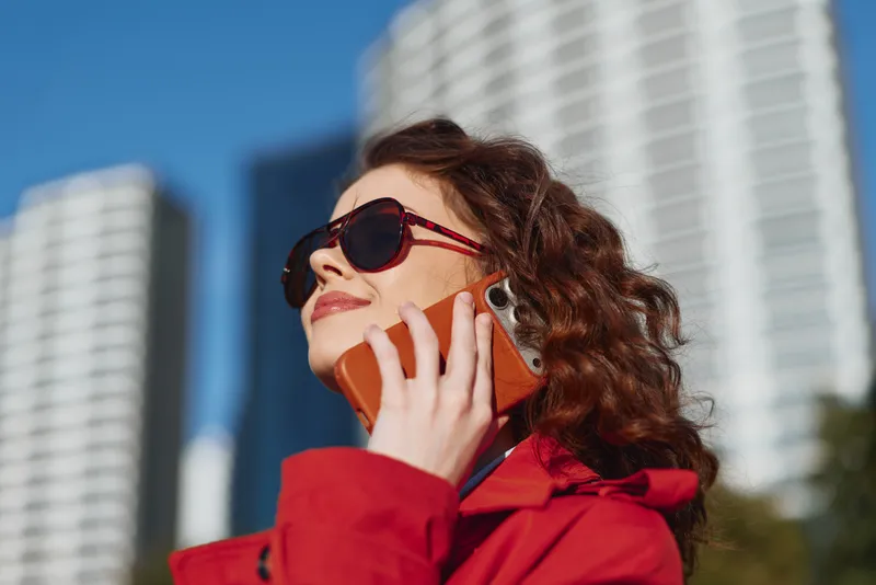 Young woman with curly hair wearing sunglasses and a red coat talks on a smartphone outdoors in an urban environment with modern buildings in the background.