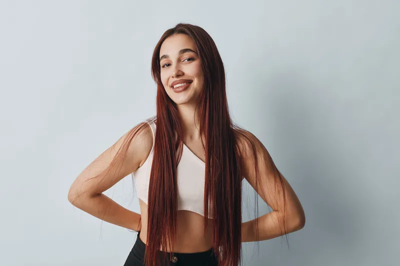 Young woman with long hair smiling and posing with hands on hips against light background. Casual outfit with sleeveless top and confident expression in studio portrait.