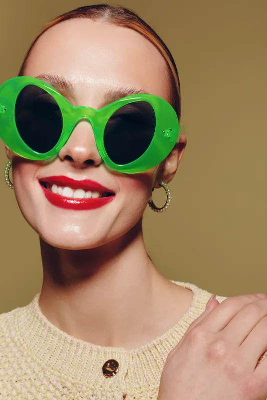 Close-up of a smiling woman wearing large green sunglasses and red lipstick. She has hoop earrings and a beige sweater, posing against a plain olive background. Fashion and style concept.