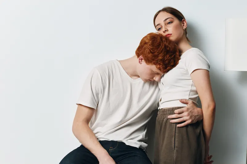 Young couple, man and woman in casual clothes in intimate pose against white background. Red hair male embracing female with white top and brown pants, relationship and connection.