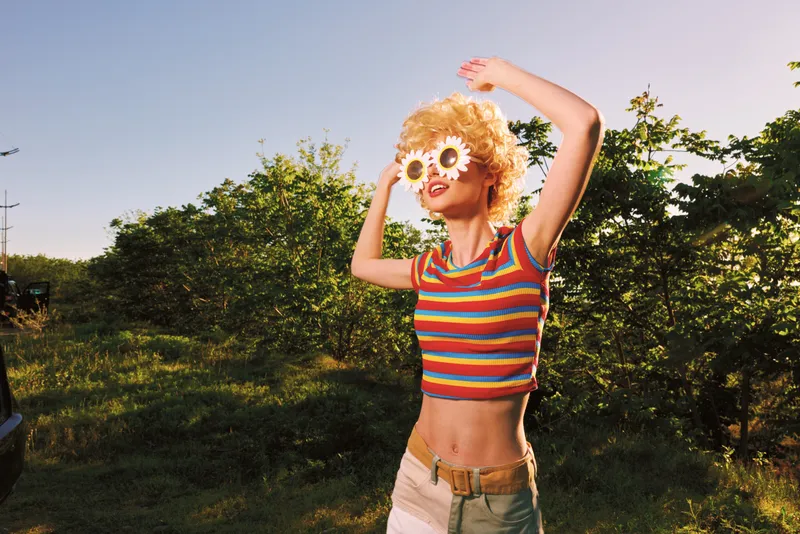 Woman enjoying sunny outdoor moment in retro film color, wearing striped crop top and light shorts, flower sunglasses, dancing arms up, carefree lifestyle, vibrant natural setting