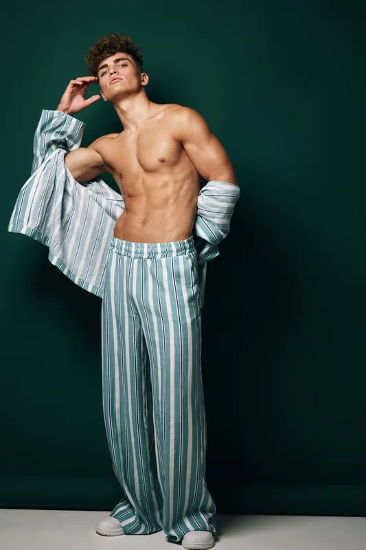 Young muscular man with curly hair wearing striped loose pajama pants and shirt open poses confidently against dark green backdrop in studio. Portrait of fit male model showing toned belly and chest