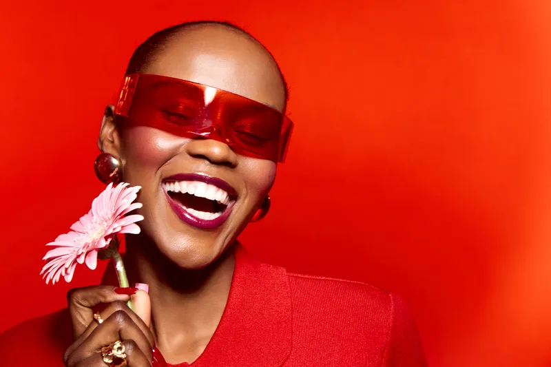 Happy African woman with dark skin wearing red sunglasses and clothing holds pink flower smiling broadly against bright red background. Concept of joy, fashion, beauty, and vibrant style.