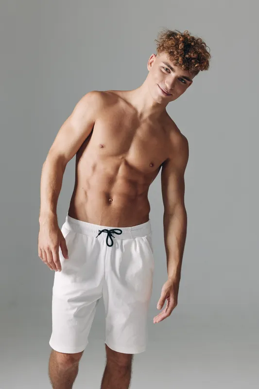 Young man with curly hair posing shirtless in white shorts against gray background, showing muscular body and confident expression in studio lighting.