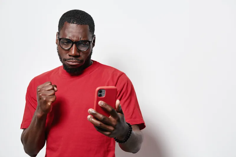 African American man with dark skin wearing glasses and red shirt looking at smartphone with clenched fist in gesture of success or excitement on white background.