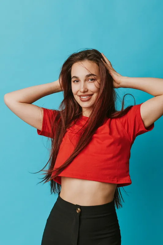 Young woman in red crop top and black pants posing with hands behind head smiling against blue background. Casual style, long hair, confident and happy expression in studio.
