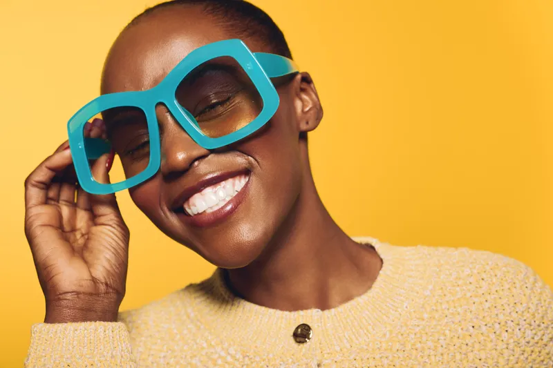 Smiling African American woman with dark skin wearing large turquoise glasses and yellow sweater poses joyfully on bright yellow background. Concept of fashion, happiness, and style.