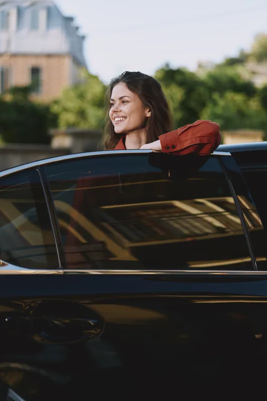 Cheerful woman in a car outdoors, smiling through the window, sunlit moment, relaxed lifestyle, scarf fluttering, urban backdrop, soft colors natural light, bright mood