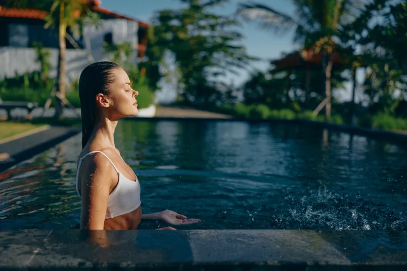 A woman stands in a bright pool at poolside, gazing toward the sun. The tropical resort scene conveys leisure and relaxation in a warm outdoor summer lifestyle.