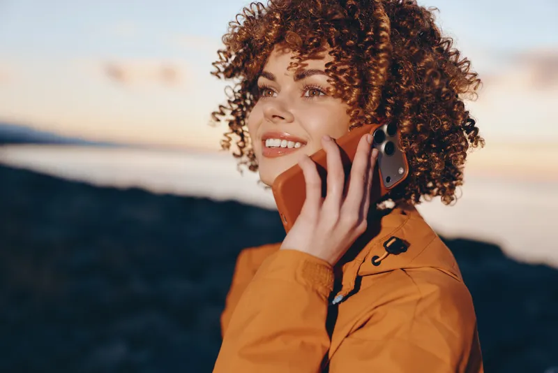 Woman lifestyle portrait, curly hair, smartphone to ear for calling, outdoors on beach coast, golden jacket in sunset daylight, calm relaxed vacation mood, with soft bokeh and sea breeze