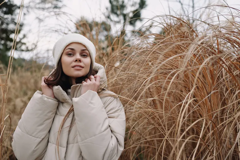 Woman in a warm coat and beanie posing outdoors among tall grasses in winter, soft natural light, calm expression, cozy fashion ambiance, seasonal outdoor portrait for lifestyle and nature inspired