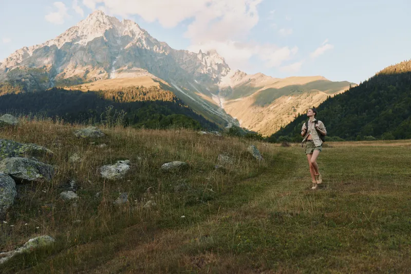 Lively woman hiking outdoors on a mountain meadow, backpack ready, sunlit trail, gentle breeze, vibrant nature, distant peaks, relaxed pose, lifestyle moment