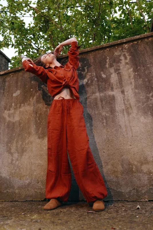 A woman in loose orange overalls strikes a bold pose against a weathered wall in an outdoor setting, conveying casual confidence and urban style in bright light