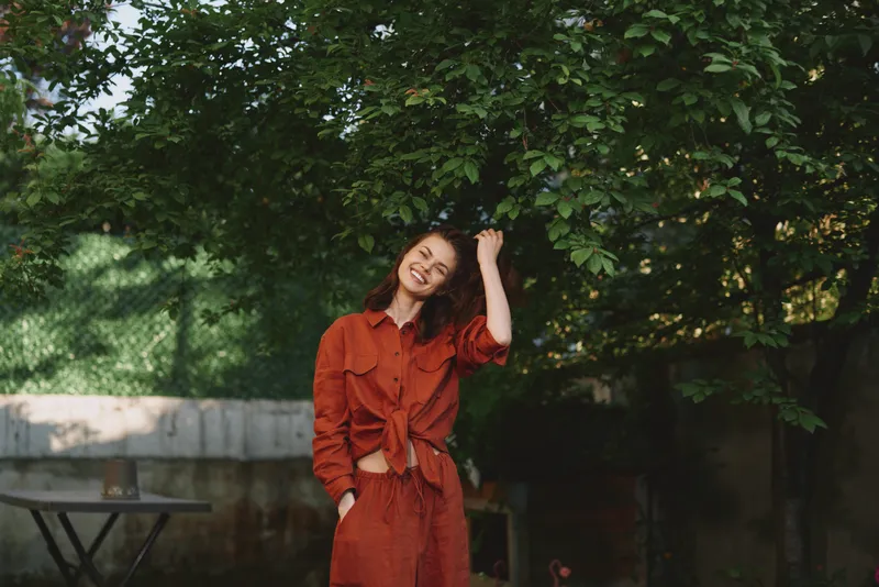 Young woman in a red outfit smiles under a lush leafy tree, posing casually in a sunlit backyard, conveying a relaxed and confident lifestyle