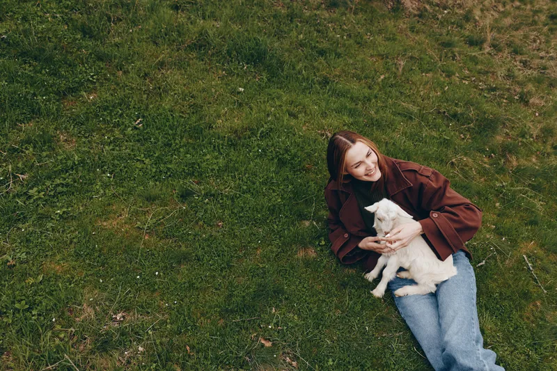 Girl lounging on grass with a goat kid in outdoors setting, showcasing lifestyle, relaxation, and affection in nature. Cozy light, casual clothing, and a peaceful mood create a warm, intimate moment