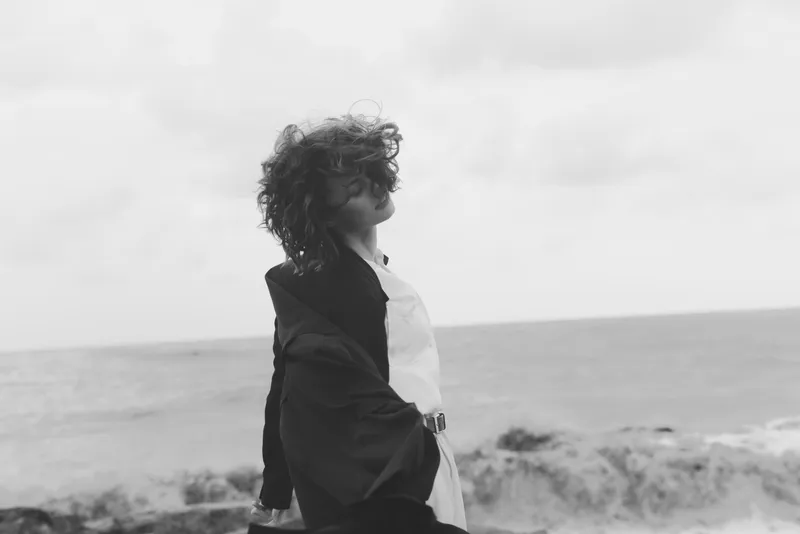 Windy sea view with a windblown woman in a loose coat, standing on the shore, gazing toward the horizon, creating a moody black and white fashion moment.