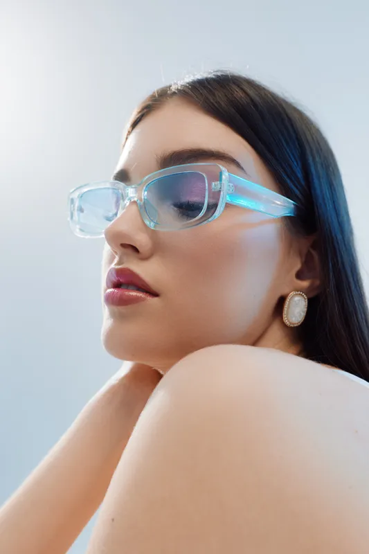 Young woman with long dark hair wearing transparent blue fashion glasses and elegant earrings poses with closed eyes. Beauty and fashion concept with soft lighting.