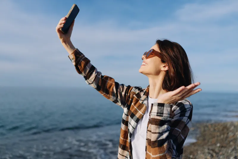 Woman selfie sea smartphone outdoor sunny beach lifestyle portrait by shore, woman captures moment with breezy seaside scenery and relaxed casual vibe