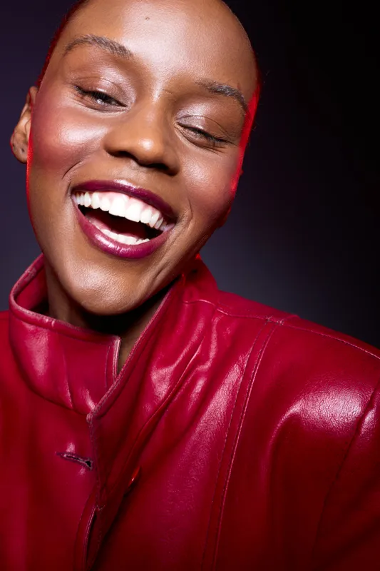 Close-up of happy African woman with dark skin laughing, wearing red leather jacket. Studio portrait with bright lighting and joyful expression. Concept of fashion, beauty, and confidence.