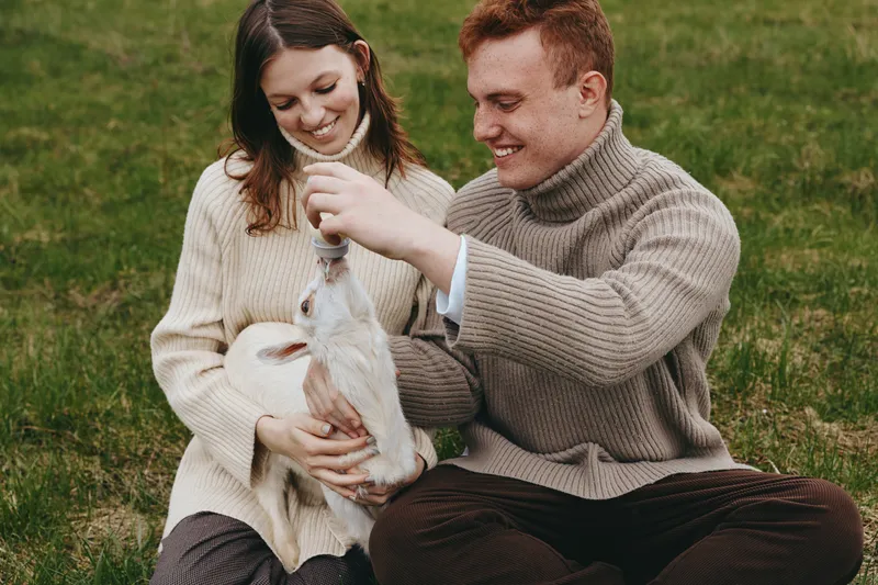 Young couple in cozy sweaters enjoys feeding a small white baby goat while sitting on green grass during a relaxed lifestyle moment outdoors in autumn.